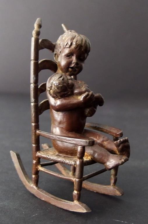 Bronze child figurine riding a rocker chair, seated and holding the steering bar; bronze figure with a raised back and side arms.