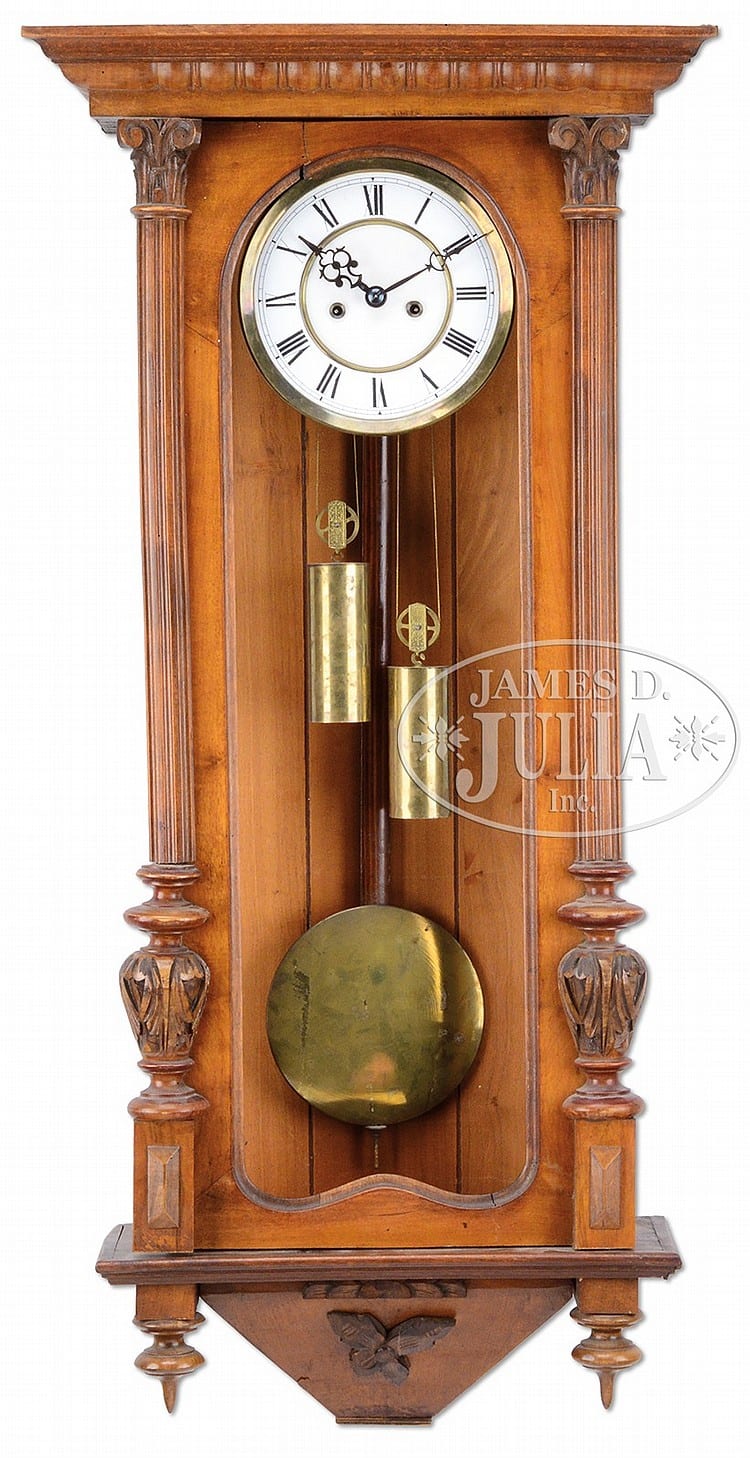 Wooden Vienna-style wall clock with an oval pendulum door, brass pendulum disk, and Roman numeral face.