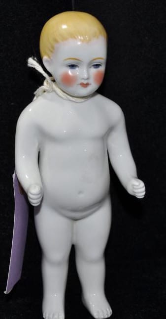 Porcelain doll figure with painted face, standing nude and holding a purple cloth or cape-like piece at the side.