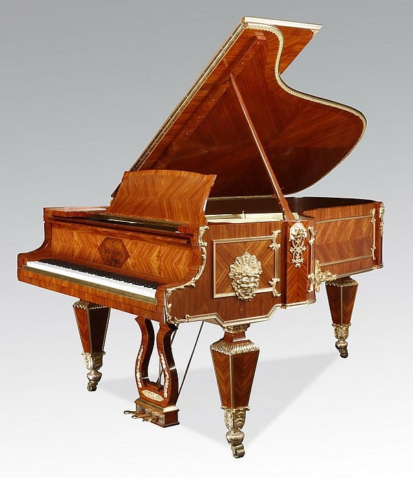 A wooden grand piano with lid open, brass trim, and ornate carved end panels on a studio gray background.