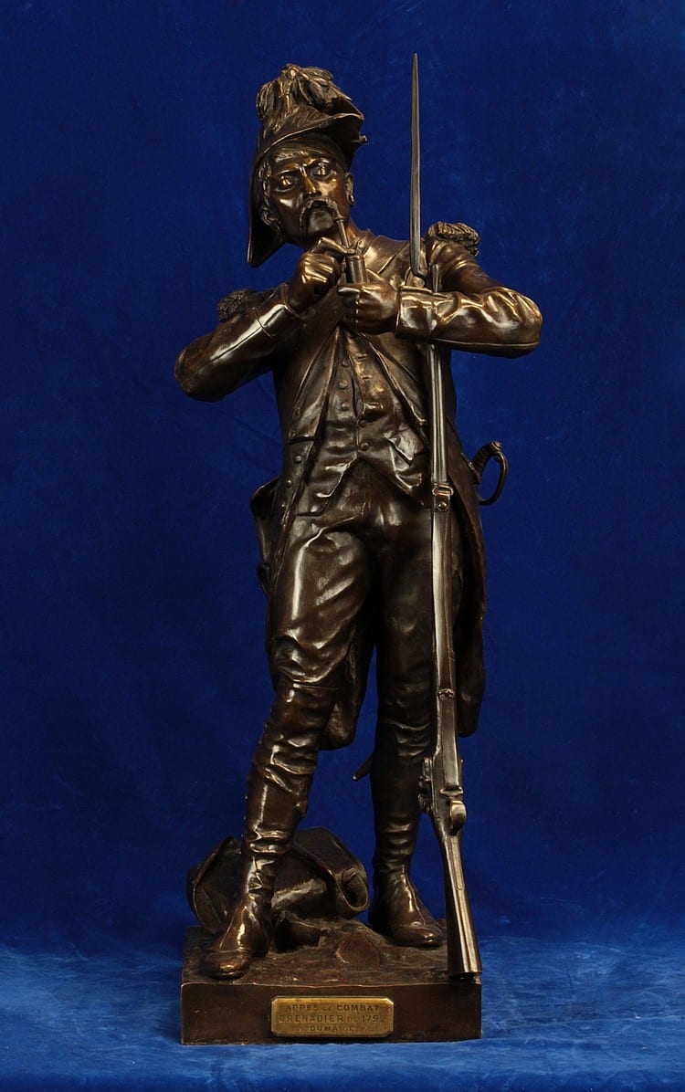 Bronze sculpture of a standing soldier holding a tall flagpole, with a raised left hand and boots on a rectangular base, against a blue background.