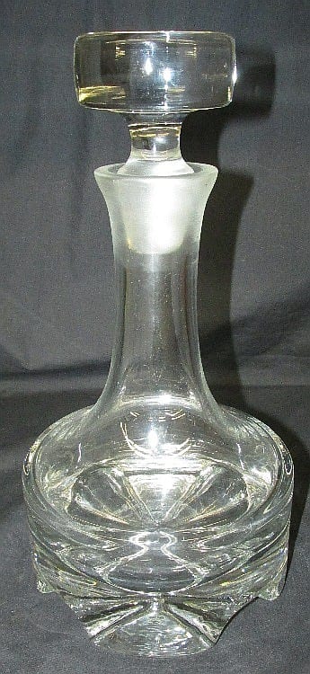 Clear glass decanter with a faceted stopper and a cut, geometric base on a dark background.
