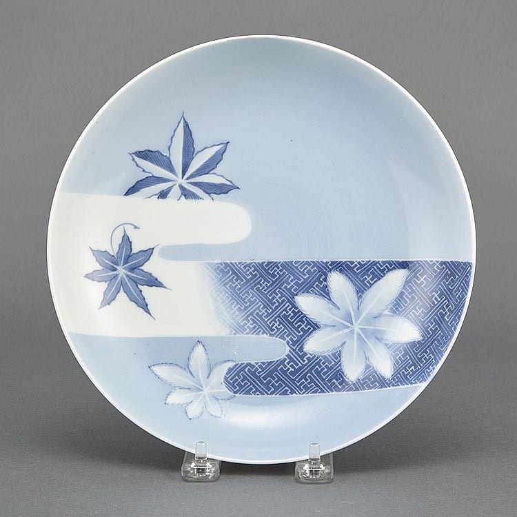 Round blue-and-white porcelain dish with four stylized maple leaf motifs and a gray geometric band, shown on a stand.