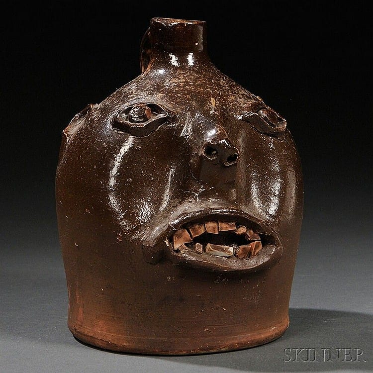 Brown-glazed stoneware face jug with a molded smiling face and open mouth, photographed on a studio background.
