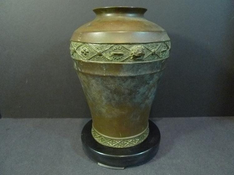 Metal vase with a wide rim and engraved banding around the neck and base, shown on a dark stand.