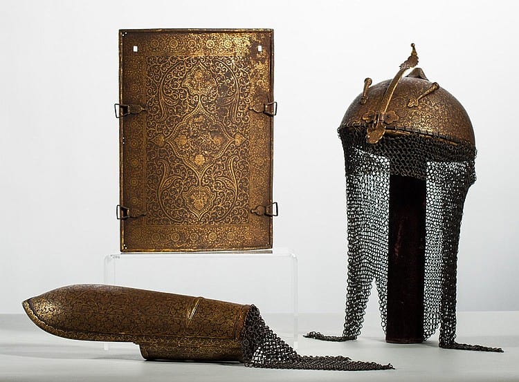 Bronze-gold helmet with chainmail curtain and a matching curved shoulder armor piece, displayed with an ornate metal panel behind it.