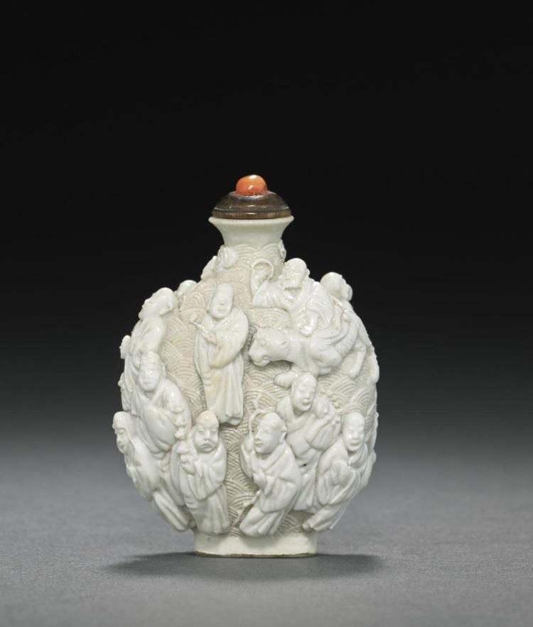 White porcelain Chinese snuff bottle with relief figures surrounding a central seated figure and a small orange stopper, shown on a gray studio background.