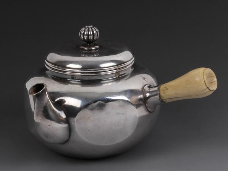Silver-colored teapot with a round knob lid and a light wood handle on a dark background.