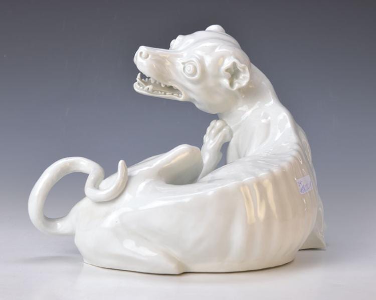 White porcelain figurine of a seated dog with an open mouth, curled tail, and a raised front paw, shown against a gray studio background.