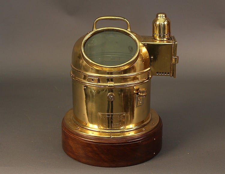 Brass binnacle compass with a round glass window, hinged metal hood, and wooden base on a dark background.