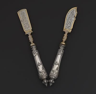 Pair of ornate fish knives with silver-toned handles and decorated blades against a dark background.