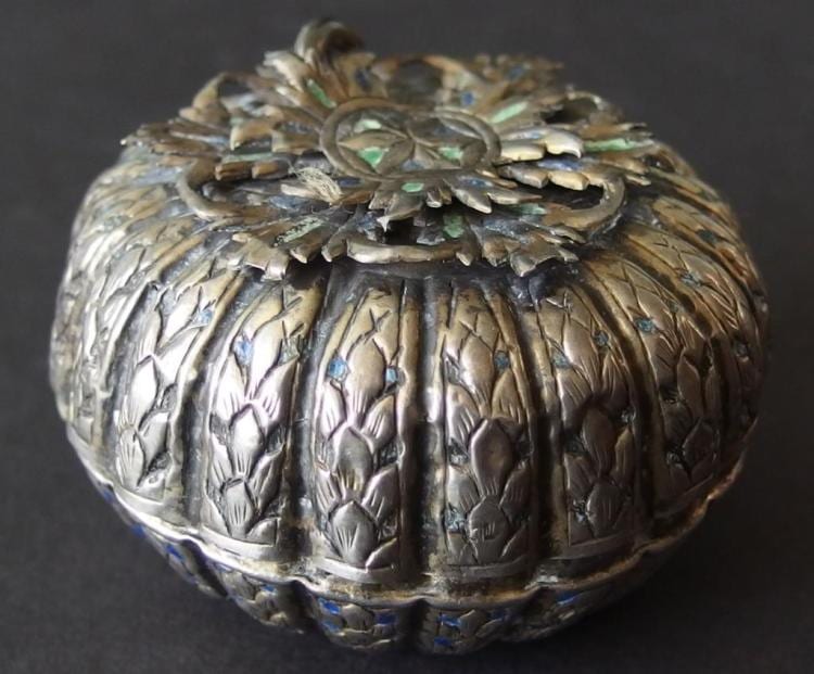 Close-up of an ornate silver-toned pill box with embossed floral pattern and a decorative hinged lid topped with a flower-like finial.