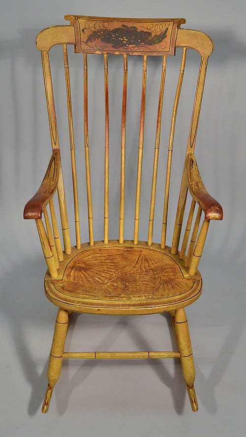 Wooden Windsor-style rocking chair with turned spindles, curved arms, and a carved painted crest on the back.