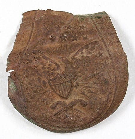 Small brown metal shield-shaped object fragment with raised decorative motifs and initials on a white background.