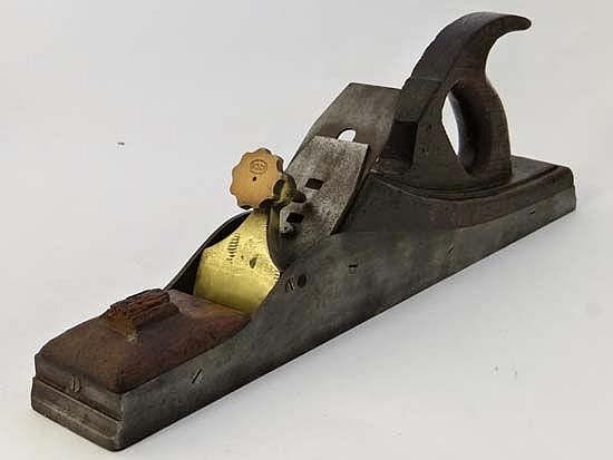 Vintage hand wood plane with a dark metal body, brass-colored front section, and a wood handle resting on a white background.
