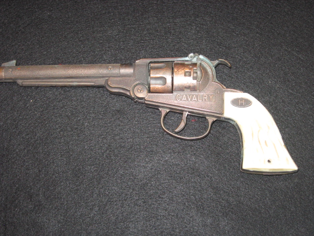 Vintage toy cap gun shaped like a revolver with a long barrel and the word “CAVALRY” on the frame, shown on a dark fabric background.