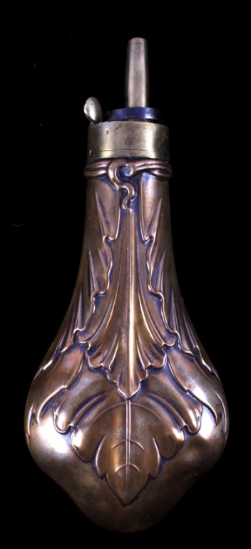 Small antique-style powder flask with brass and copper color and engraved floral scrollwork, shown against a black background with a metal cap on top.
