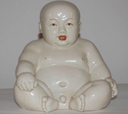 White porcelain figure of a seated baby with a bald head, rosy lips, and hands resting forward against a plain background.