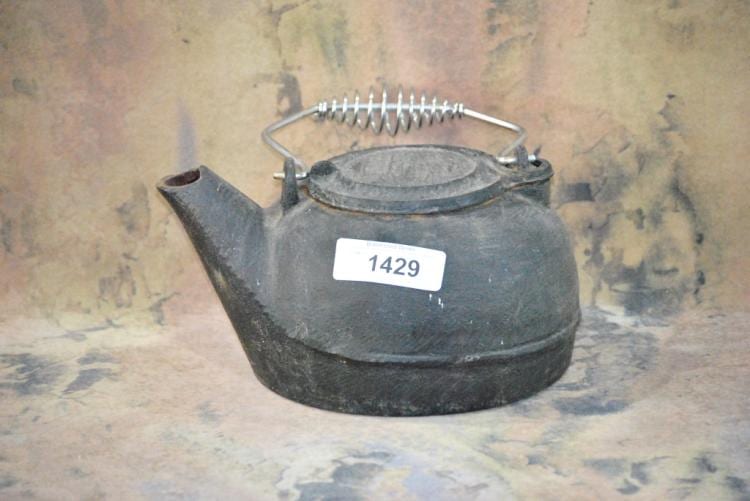 Unmarked black cast iron tea kettle with curved handle, spout, and wire handle rest, photographed on a mottled background.