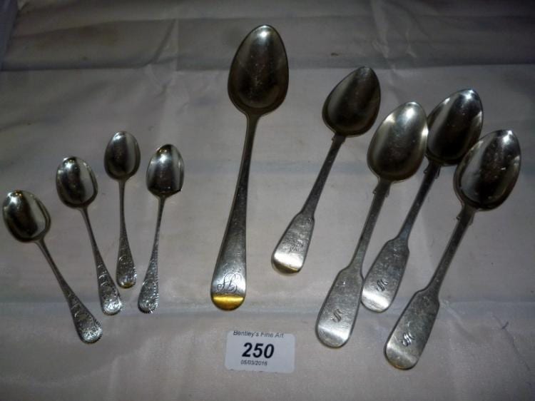 Group of silver teaspoons and dessert spoons with engraved handles, photographed on a neutral fabric background.
