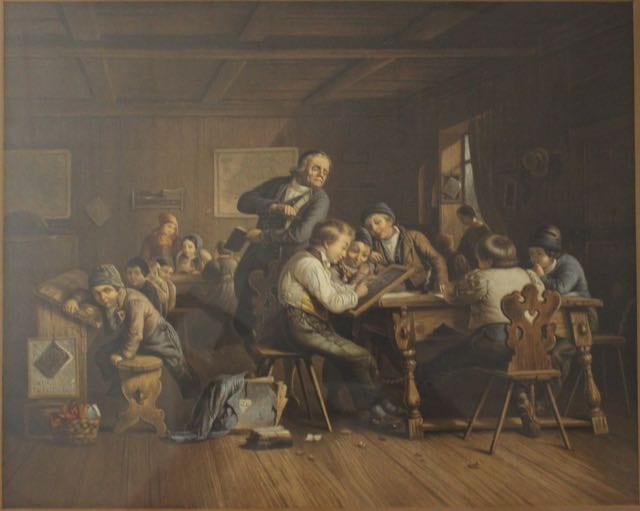 A group of children and adults gathered at a table and desks inside a classroom or music room, with one man standing and teaching.