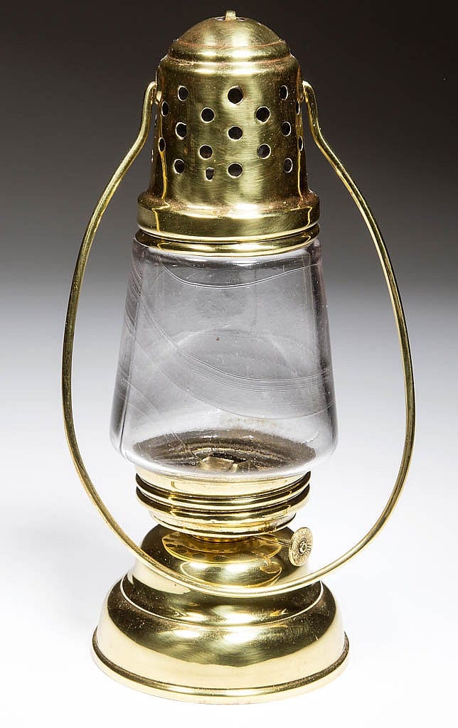 Brass hurricane lamp with perforated burner cap and clear glass chimney, shown against a dark background.