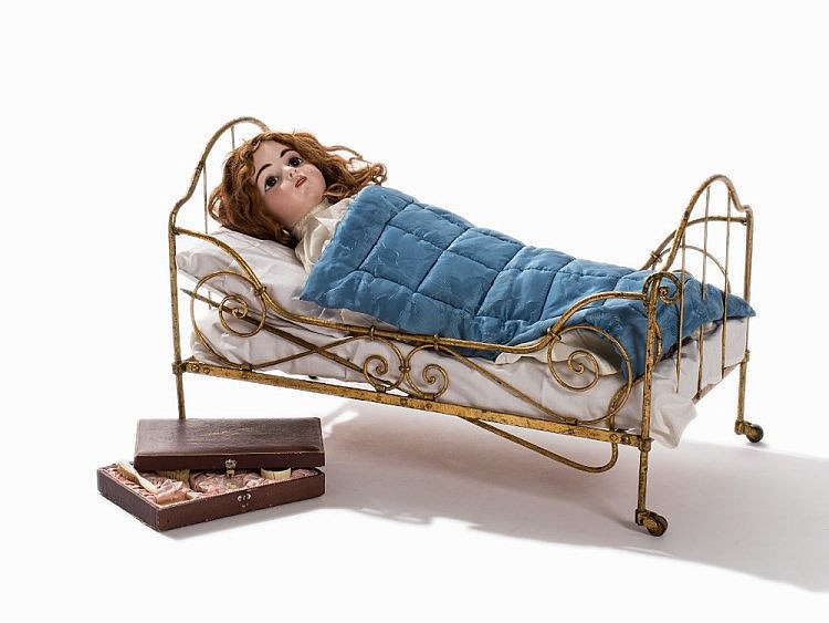 Porcelain-style doll lying in a metal-framed bed with a blue quilt and white pillows, with a small box beside it.