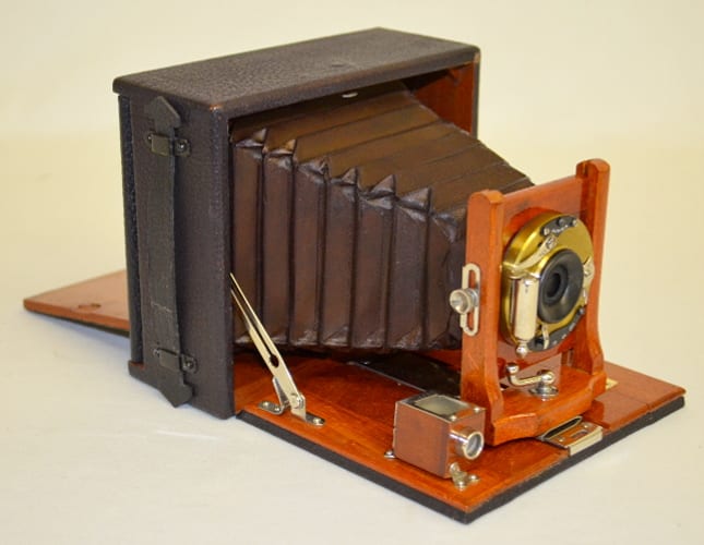 Vintage bellows box camera shown open, with wooden body, accordion bellows, and front lens panel on a base.