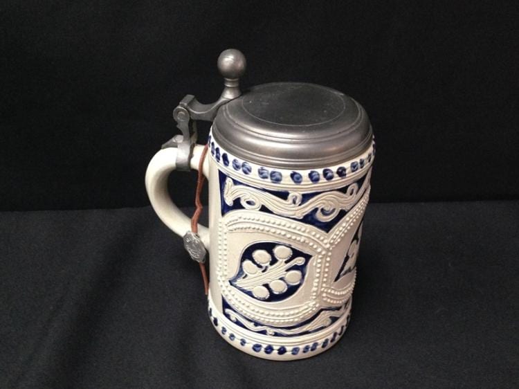 Glazed ceramic beer stein with blue and white bands and an embossed crest, metal lid and handle, shown against a dark background.