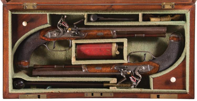 Open wooden case displaying two antique-style dueling pistols with carved grips and metal barrels, plus ammunition accessories in compartments.