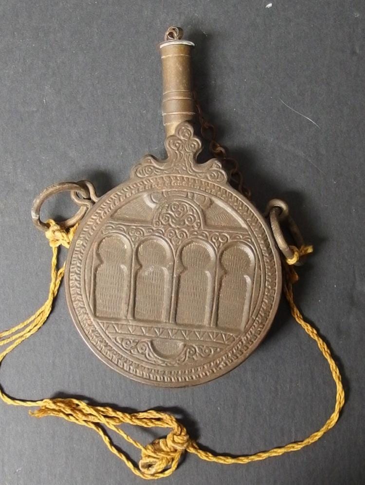 Antique-style metal powder flask with a rounded front panel, decorative reliefs, and a loop handle, shown on a dark background.
