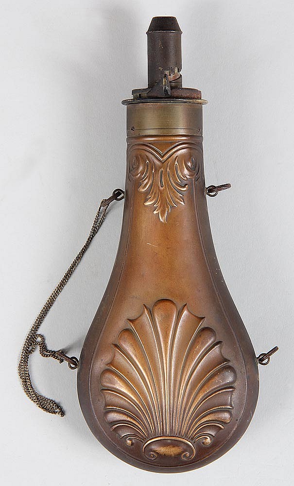 Antique-style metal black powder flask with a hinged top and engraved leaf design on the front, shown upright with side rings and a chain.