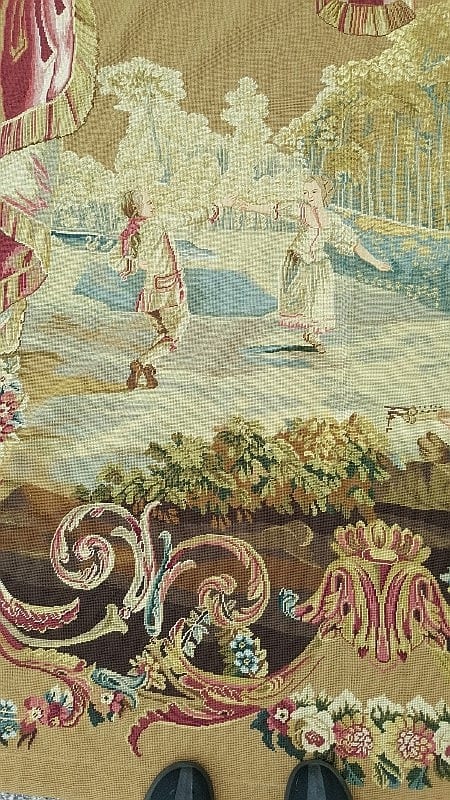 Close-up of a stitched needlepoint tapestry showing a pastoral scene with people near a river or landscape, with floral ornaments below.