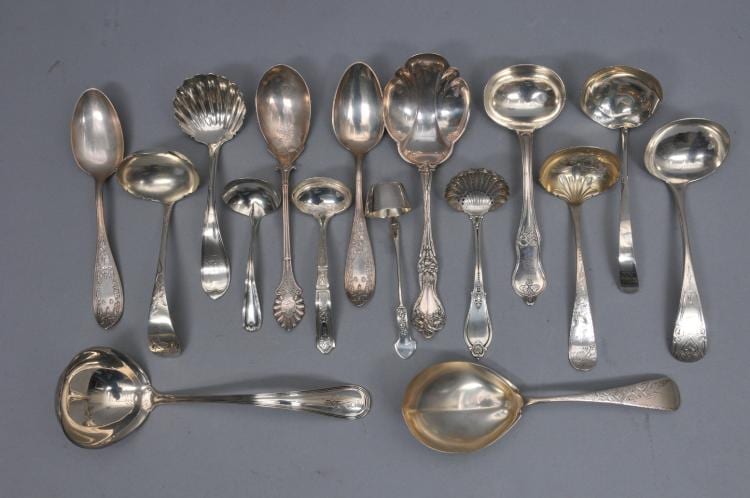 Assorted polished silver-colored serving spoons and ladles laid out side by side against a gray background.