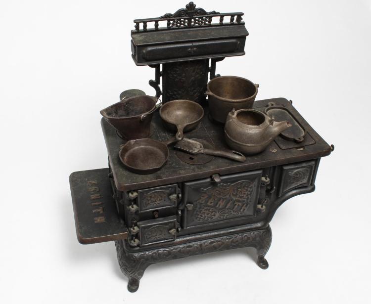 Cast iron stove with ornate panels and several small metal pots and cups arranged on top, shown against a white background.