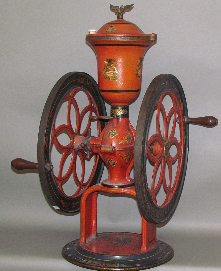 Red and orange hand-crank coffee grinder with a decorative finial on top and large spoked wheels, shown on a stand.