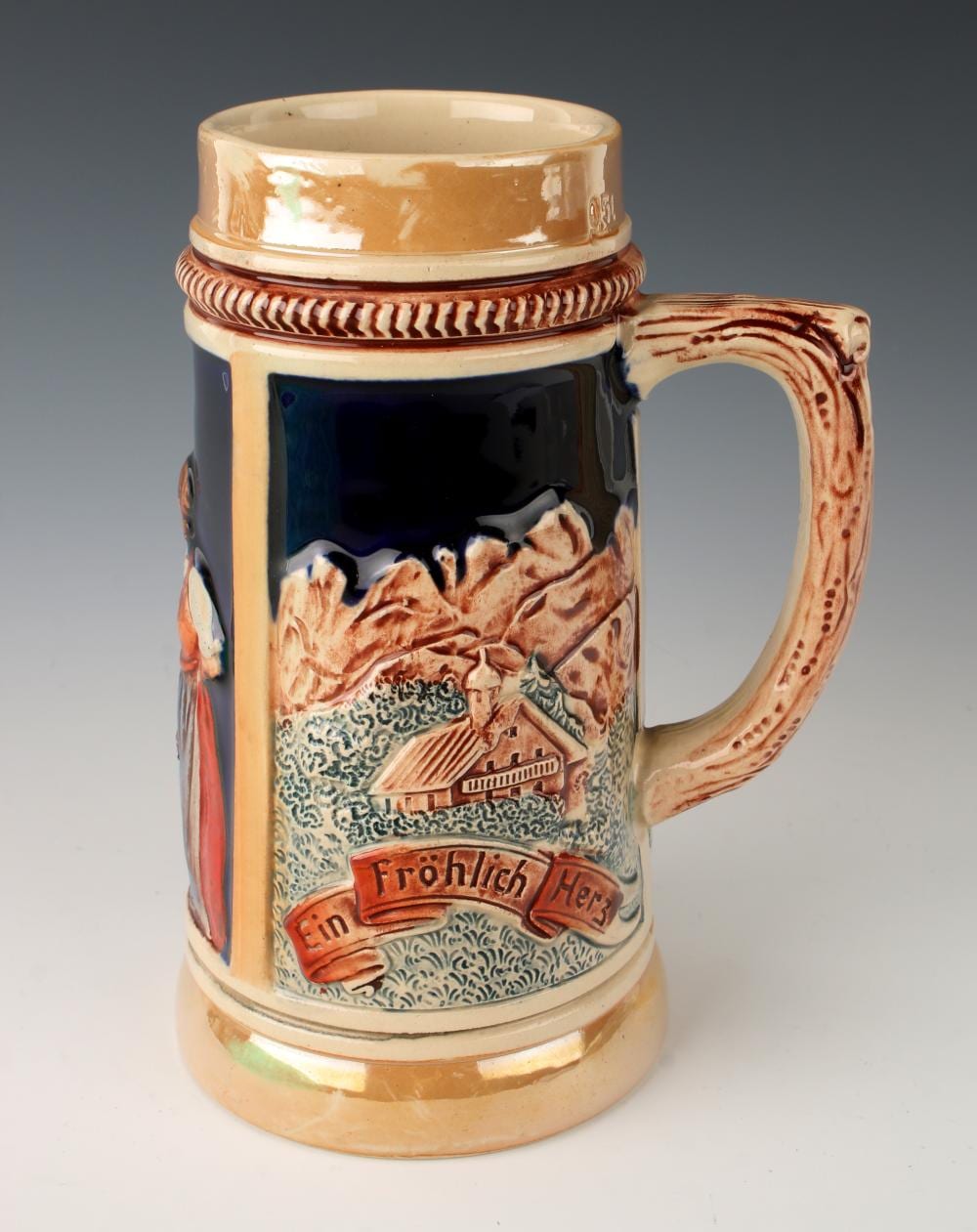 German beer stein with a blue and brown painted scene and red banner reading “Fröhlich Herr,” with a strap handle and ribbed rim.