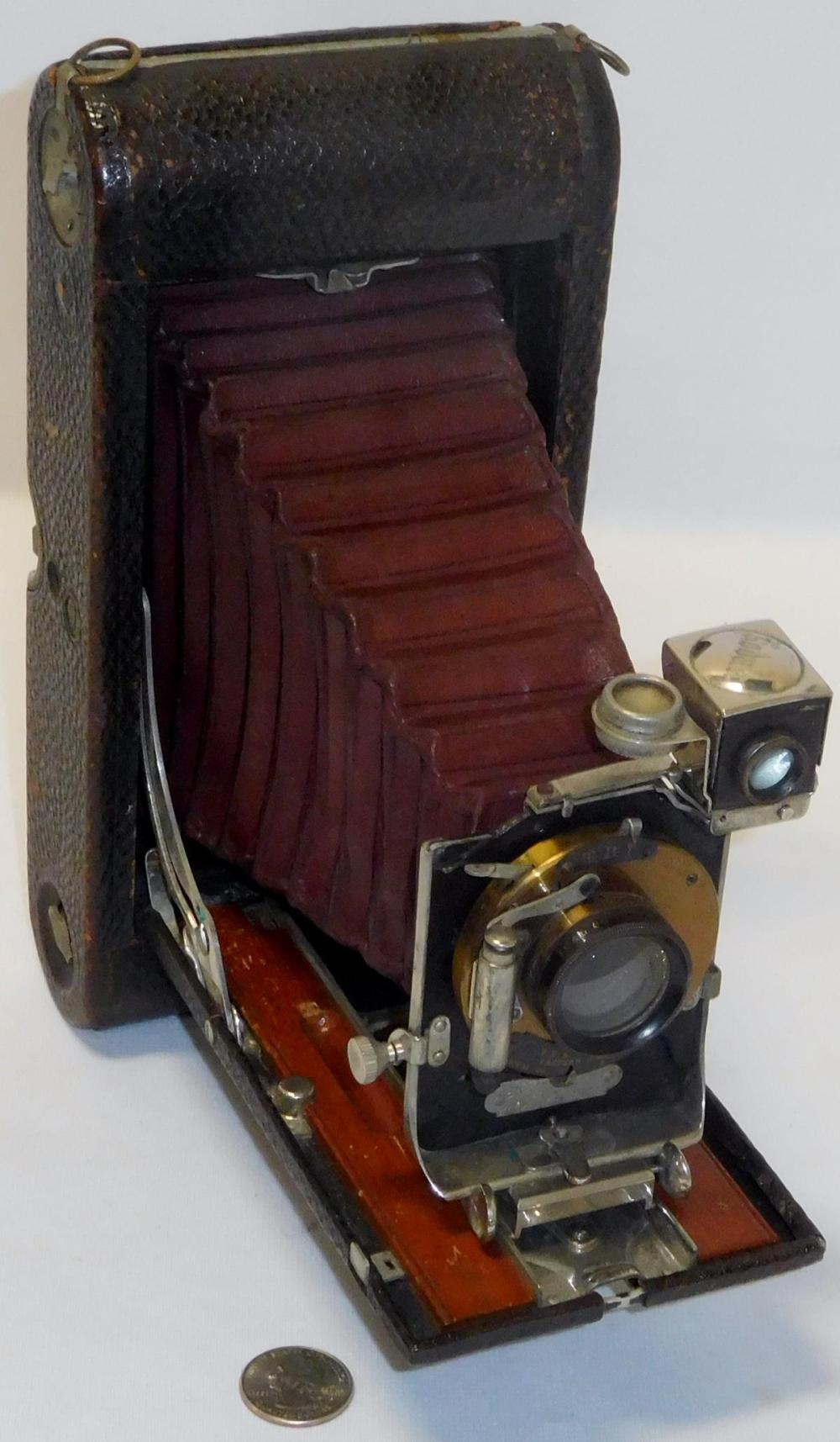 Close-up of an antique folding camera with a black body, red bellows, and a visible lens assembly, shown open on a white background.