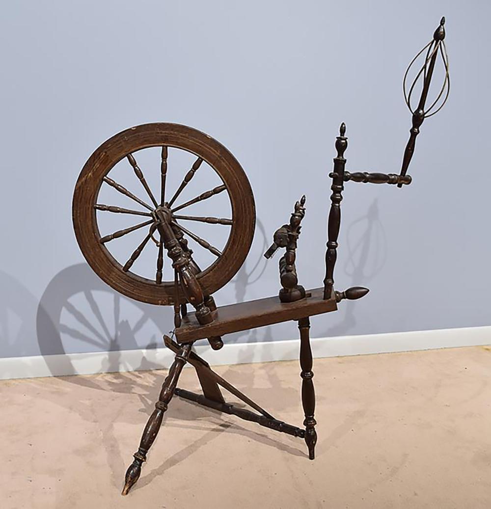 Wooden spinning wheel with a round wheel and turned spindle supports on a beige floor against a light gray background.