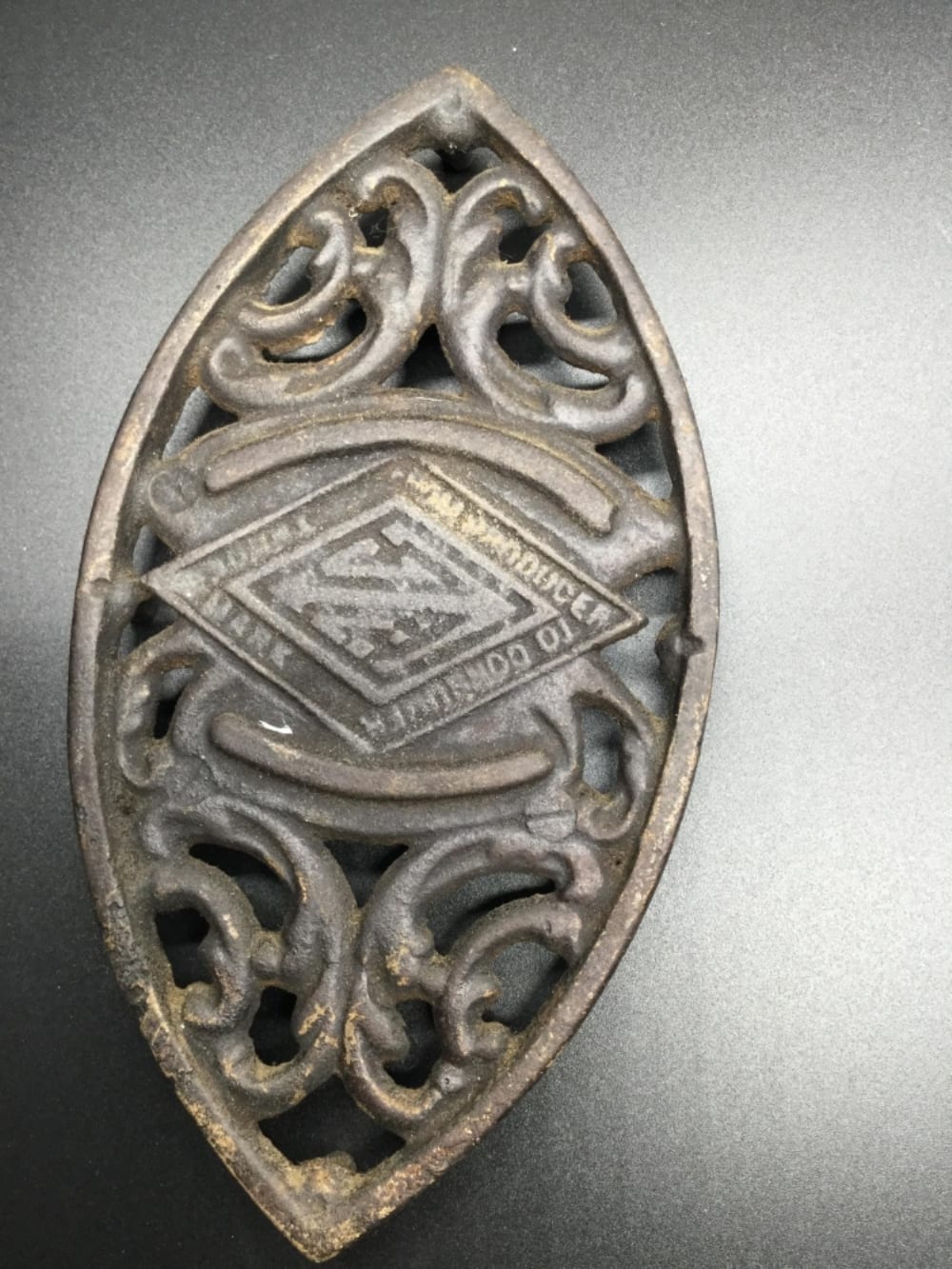 Cast iron trivet with decorative scrollwork and a central diamond-shaped emblem, shown in close-up against a gray background.