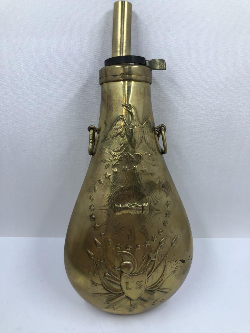 Antique-style brass powder flask with a vertical spout, hinged cap, side handles, and engraved stars and an eagle design on the front.