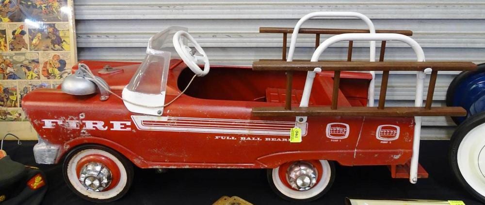 Red fire truck pedal car with white steering wheel and handrails, shown side view indoors.