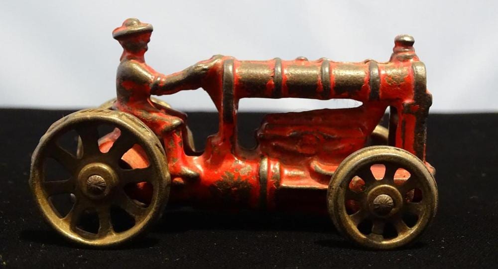 Small red and gold cast-iron farm tractor toy with four-spoke wheels and a raised exhaust stack on a dark surface.