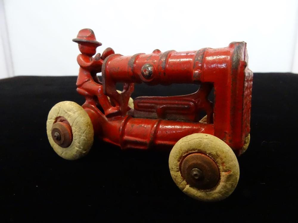 Red cast-iron farm tractor toy with four wheels and a seated driver figure, photographed on a dark background.