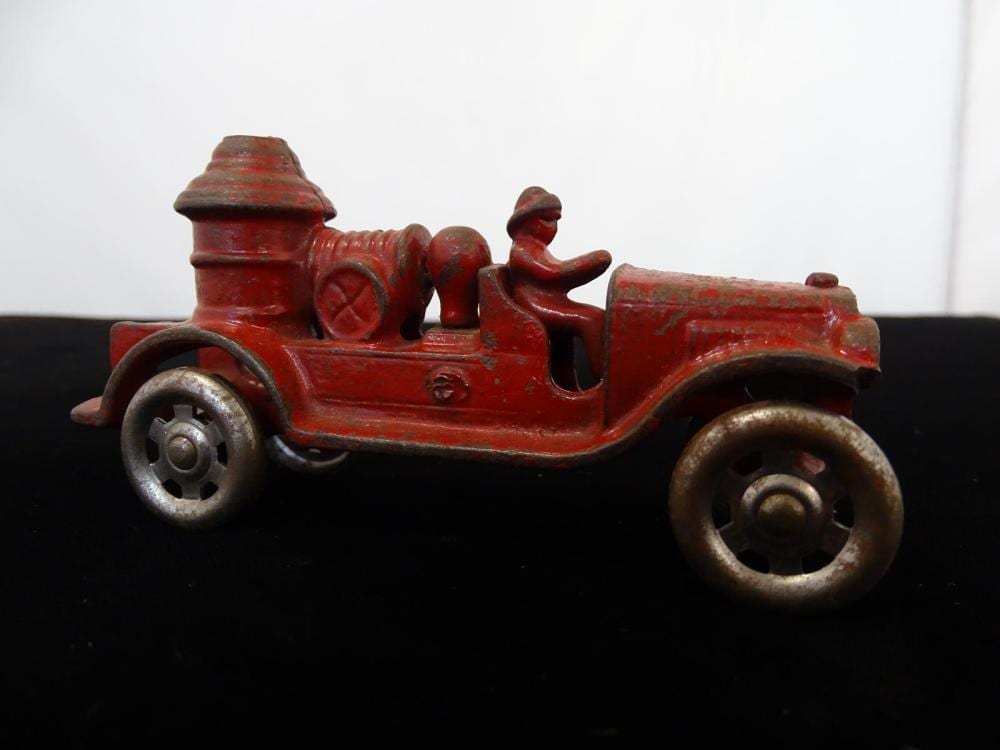 Red cast-iron toy fire pumper truck with detailed cab, wheels, and front nozzle, shown in profile against a black background.