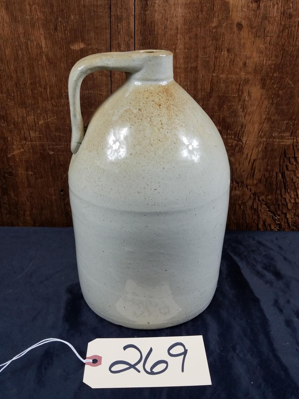 White speckled stoneware jug with a curved handle on a dark cloth, photographed against a wooden wall.