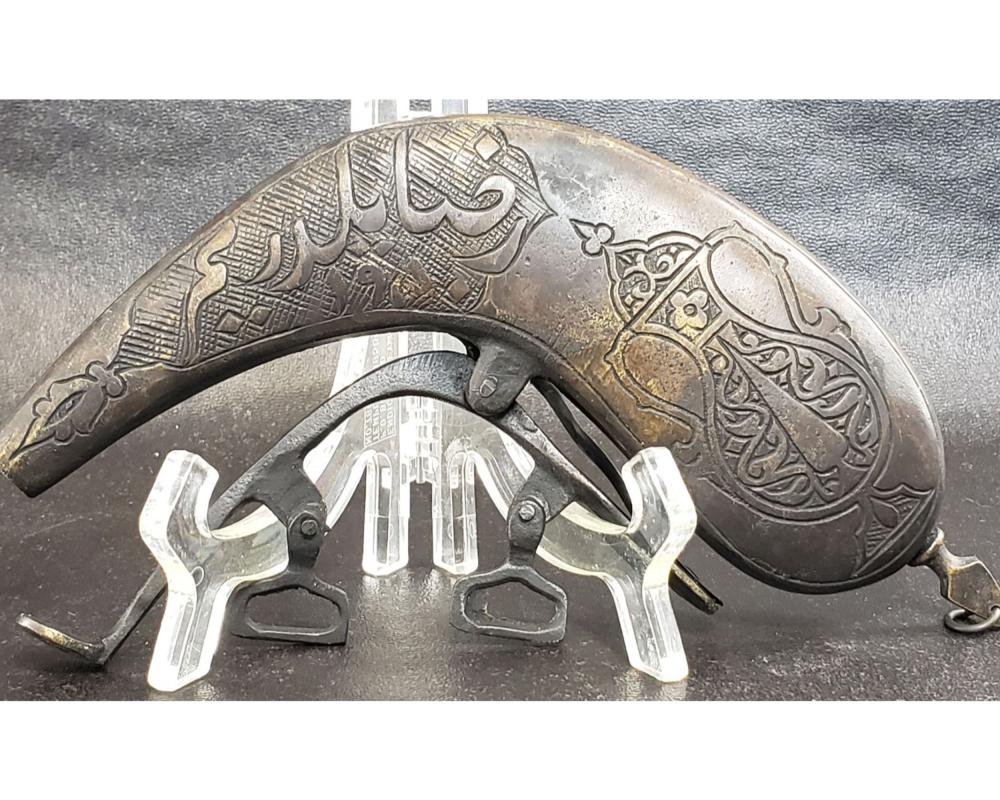 Antique-style metal powder flask with engraved Islamic-style calligraphic designs and a black powder measure stand.