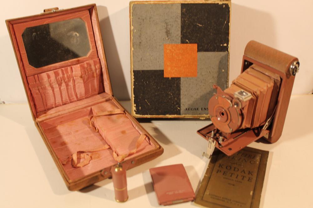 Vintage Kodak camera with a brown case open, showing camera body, booklet, and a separate Kodak box and camera lens assembly.