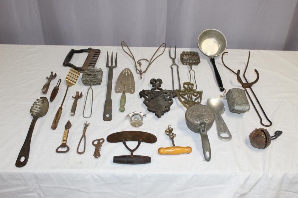 Assorted vintage kitchen utensils and cookware tools laid out on a white tablecloth, including a whisk, ladle, grater, and various metal gadgets.