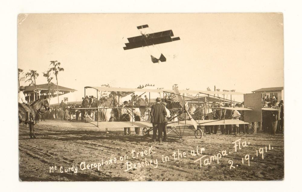 Sepia real-photo postcard showing an early aircraft on a grassy airfield with people gathered near tents and equipment; sky with plane overhead.